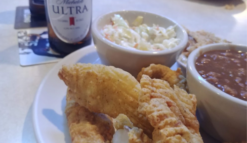 A plate of fried fish, coleslaw, and baked beans, with a bottle of beer in the background.