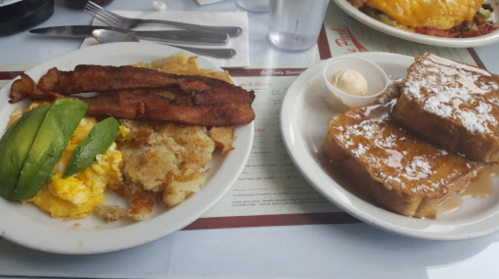 A plate of breakfast featuring scrambled eggs, bacon, hash browns, avocado, and French toast with syrup and butter.