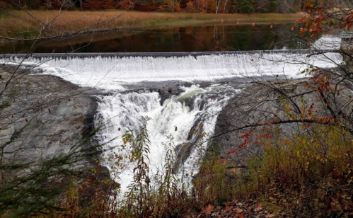 A waterfall cascading over rocks into a calm river, surrounded by autumn foliage and a serene landscape.