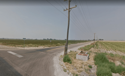 A rural road intersects with a field, featuring power lines and a concrete structure near the roadside.
