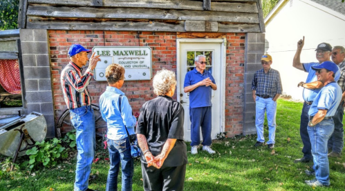 A group of people gathered outside a small building, discussing and pointing at a sign that reads "Lee Maxwell, Collector of the Unusual."