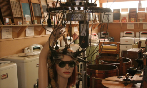 A vintage mannequin with sunglasses and wires on its head, surrounded by old appliances in a museum-like setting.
