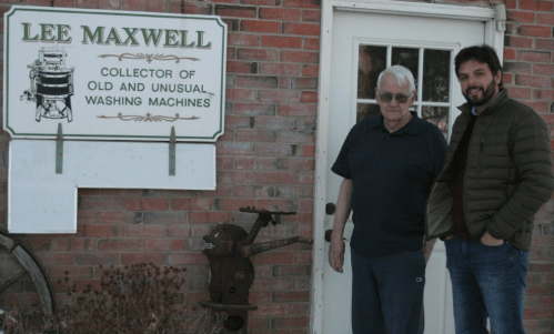 Two men stand beside a sign that reads "Lee Maxwell, Collector of Old and Unusual Washing Machines," in front of a brick wall.