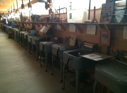 A row of vintage washing machines displayed in a museum, with various artifacts and signs on the wall above.
