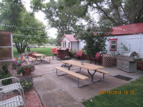 A cozy outdoor space with picnic tables, a grill, and small cabins, surrounded by greenery and string lights.