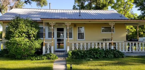 A charming yellow house with a metal roof, surrounded by greenery and a small porch with white railings.