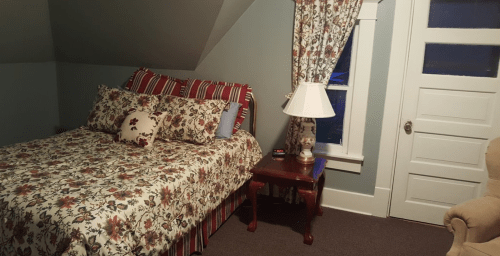 Cozy bedroom with a floral bedspread, bedside table, lamp, and patterned curtains in a softly lit space.