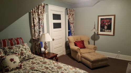 Cozy bedroom with floral bedding, a beige armchair, lamp, and floral curtains, featuring a door and wall art.