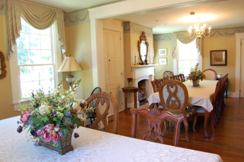 A bright dining room with a large table, elegant chairs, floral centerpiece, and a cozy fireplace.