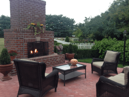 Cozy outdoor seating area with a brick fireplace, a pumpkin, and lush greenery in the background.
