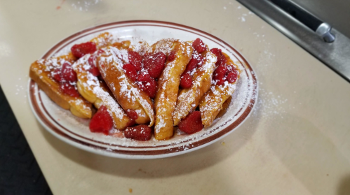 A plate of golden-brown French toast sticks topped with fresh raspberries and dusted with powdered sugar.