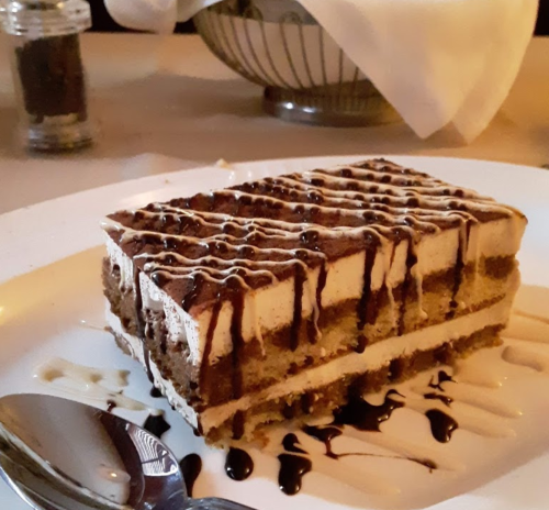 A slice of layered dessert with coffee-flavored cream, drizzled with chocolate sauce, on a white plate.