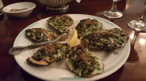 A plate of baked oysters topped with green herbs and a lemon wedge, served in a restaurant setting.