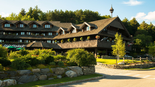 A charming lodge with a wooden exterior, surrounded by greenery and mountains, under a clear blue sky.