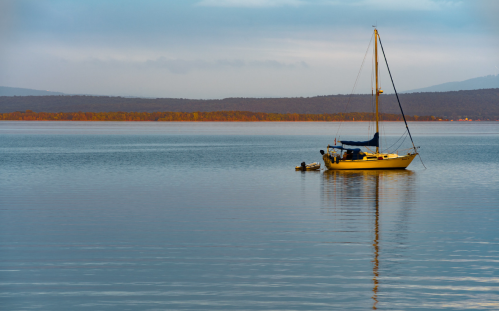 A calm lake at dawn with a sailboat anchored, reflecting on the water's surface and surrounded by distant hills.