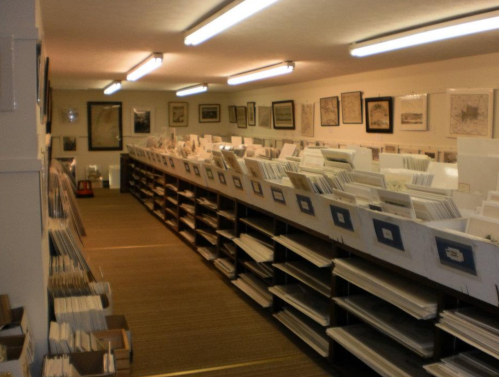 A long aisle in a storage room filled with neatly organized boxes and shelves of documents and framed pictures.