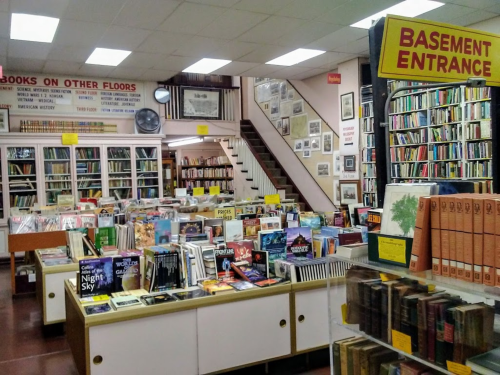 A cozy bookstore interior with shelves of books, a staircase, and a sign for the basement entrance.