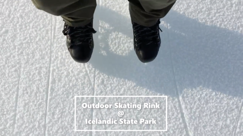 Person's feet in black skates on an outdoor ice rink at Icelandic State Park, with snow-covered ground.