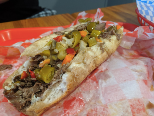 A loaded sandwich with sliced beef, pickles, and peppers on a toasted hoagie roll, served on a red tray.