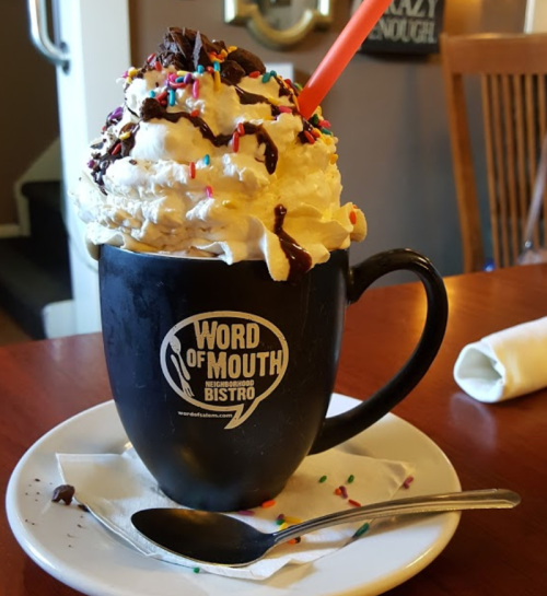 A large dessert drink topped with whipped cream, chocolate, and sprinkles in a black mug on a plate.