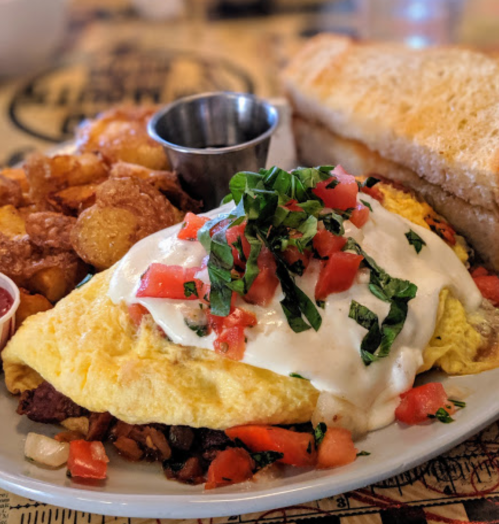 A hearty breakfast plate featuring an omelet topped with tomatoes and herbs, crispy potatoes, and a side of toast.