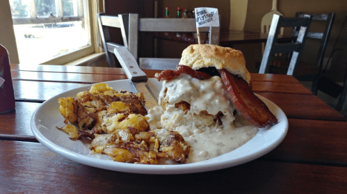 A hearty breakfast plate featuring a biscuit sandwich with bacon and gravy, alongside crispy potatoes.