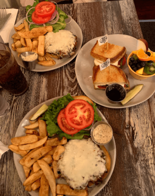 Two plates of food featuring burgers with cheese, fries, and a side of salad, plus a plate of sandwiches and fruit.