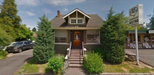 A charming green bistro with a front porch, surrounded by trees and flowers, on a sunny day.