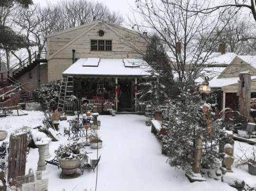 A snowy scene of a rustic building surrounded by winter decorations and garden ornaments.