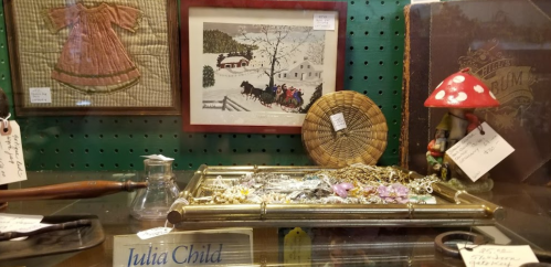 A display case featuring vintage jewelry, a framed winter scene, a woven basket, and a decorative mushroom.