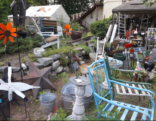A cluttered garden filled with various items, including chairs, planters, and decorative flowers among rocks and greenery.