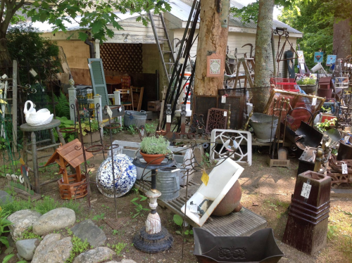 A collection of vintage items and garden decor displayed outdoors among trees and a building.