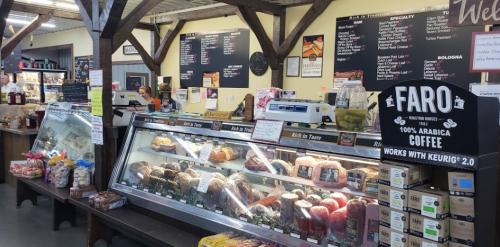 A deli counter displaying various meats and cheeses, with a coffee sign and menu boards in a market setting.