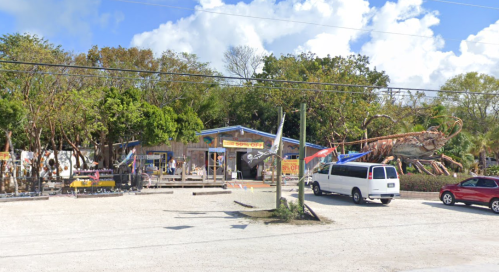 A colorful seafood shop with a large lobster statue outside, surrounded by trees and parked cars.