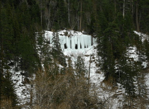 A snowy hillside with tall evergreen trees and a frozen ice formation in the background.