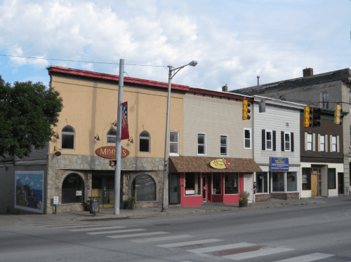 A row of small businesses on a street, featuring varied architectural styles and signage, under a cloudy sky.