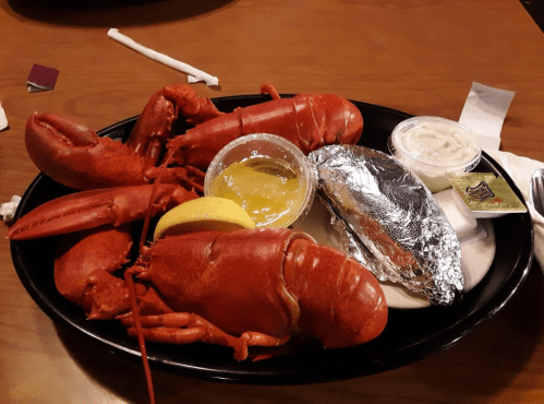 A plate with two cooked lobsters, a baked potato wrapped in foil, lemon, and dipping sauces.