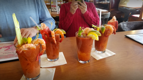 Four colorful cocktails with shrimp, lime, and celery on a wooden table, with a person in a red sweater in the background.