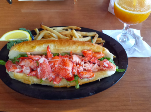 A lobster roll filled with lobster meat on a bun, served with fries and a drink on the side.