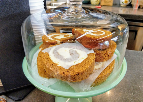 A glass cake dome displays slices of pumpkin roll cake on a green pedestal.