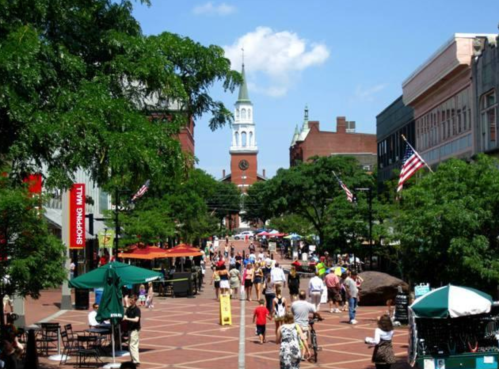 A bustling street scene with people walking, shops, and a clock tower in the background under a clear blue sky.