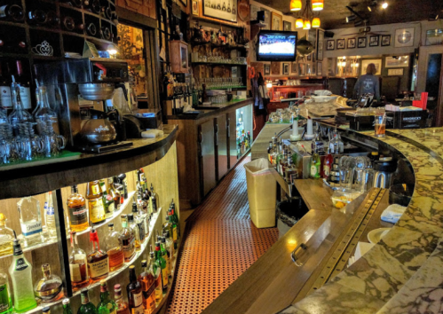 A cozy bar interior featuring a curved counter, shelves of liquor, and a TV displaying a sports game.