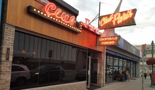 Exterior of Club Paris restaurant with neon signs, large windows, and outdoor seating on a cloudy day.