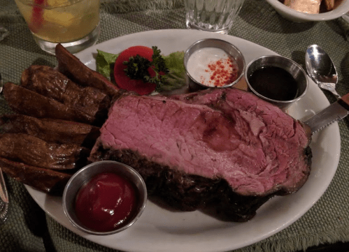 A plate featuring a large slice of prime rib, baked potatoes, and various sauces, garnished with lettuce and tomato.