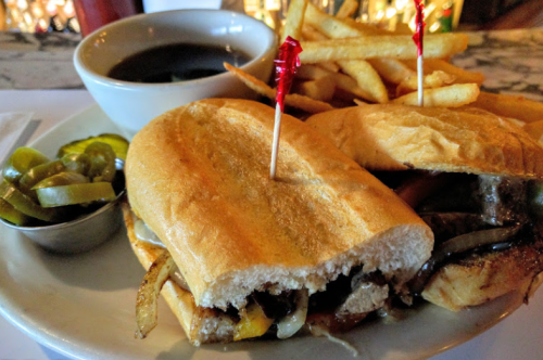 A sandwich with grilled meat, onions, and cheese, served with fries, pickles, and a side of dipping sauce.