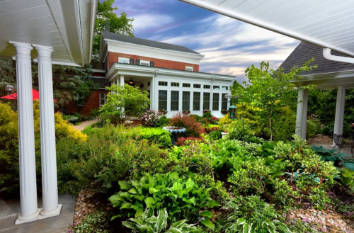 A lush garden with various plants surrounds a brick house, under a cloudy sky. Columns frame the view.