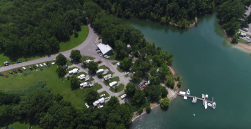 Aerial view of a lakeside campground with RVs, trees, and a small dock along the water's edge.