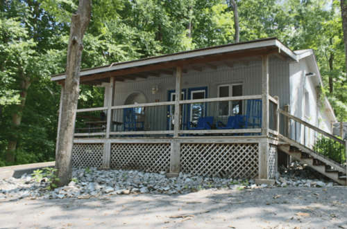 A cozy cabin with a porch, surrounded by trees and gravel, set in a serene outdoor environment.