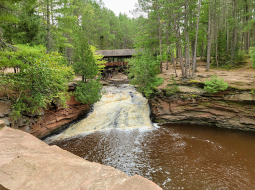 A serene waterfall flows into a calm river, surrounded by lush green trees and a rustic wooden bridge in the background.