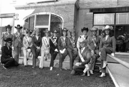 A group of pageant contestants in cowboy hats and sashes pose together outside a building.